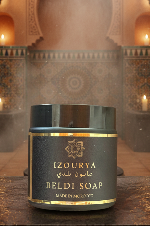 A jar of IZOURYA BELDI SOAP sits on a dark wooden surface in front of a softly blurred Moroccan-style bathroom or spa interior with arched walls, hanging lanterns, and candles. The jar is dark gray with gold lettering and a gold band, with a subtle mist or steam around it.