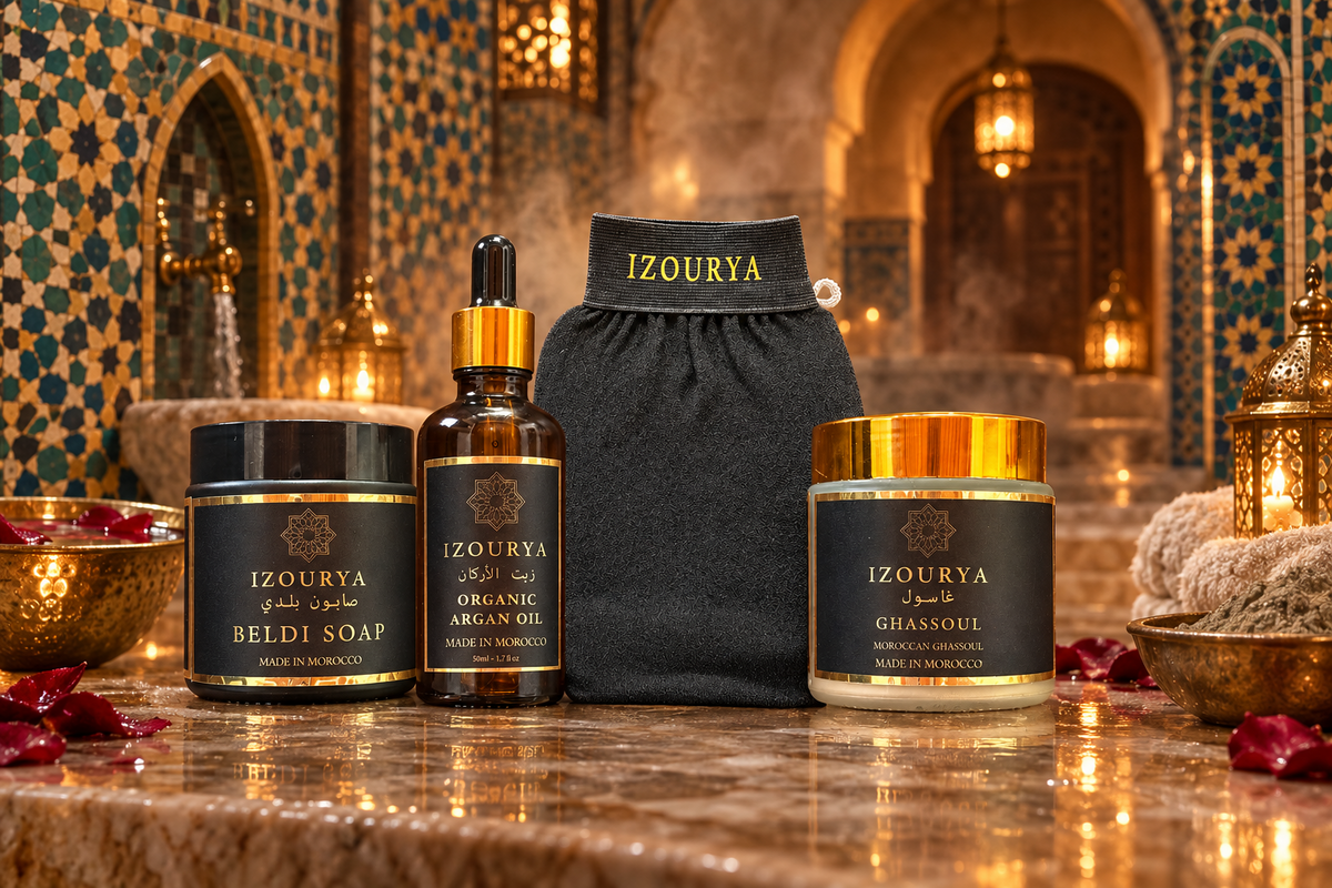 A set of three skincare products from the brand IZOURYA, including a black soap jar, an organic argan oil dropper bottle, and a ghassoul powder jar, arranged symmetrically on either side of a kessa glove. The products feature black and gold labels and caps,