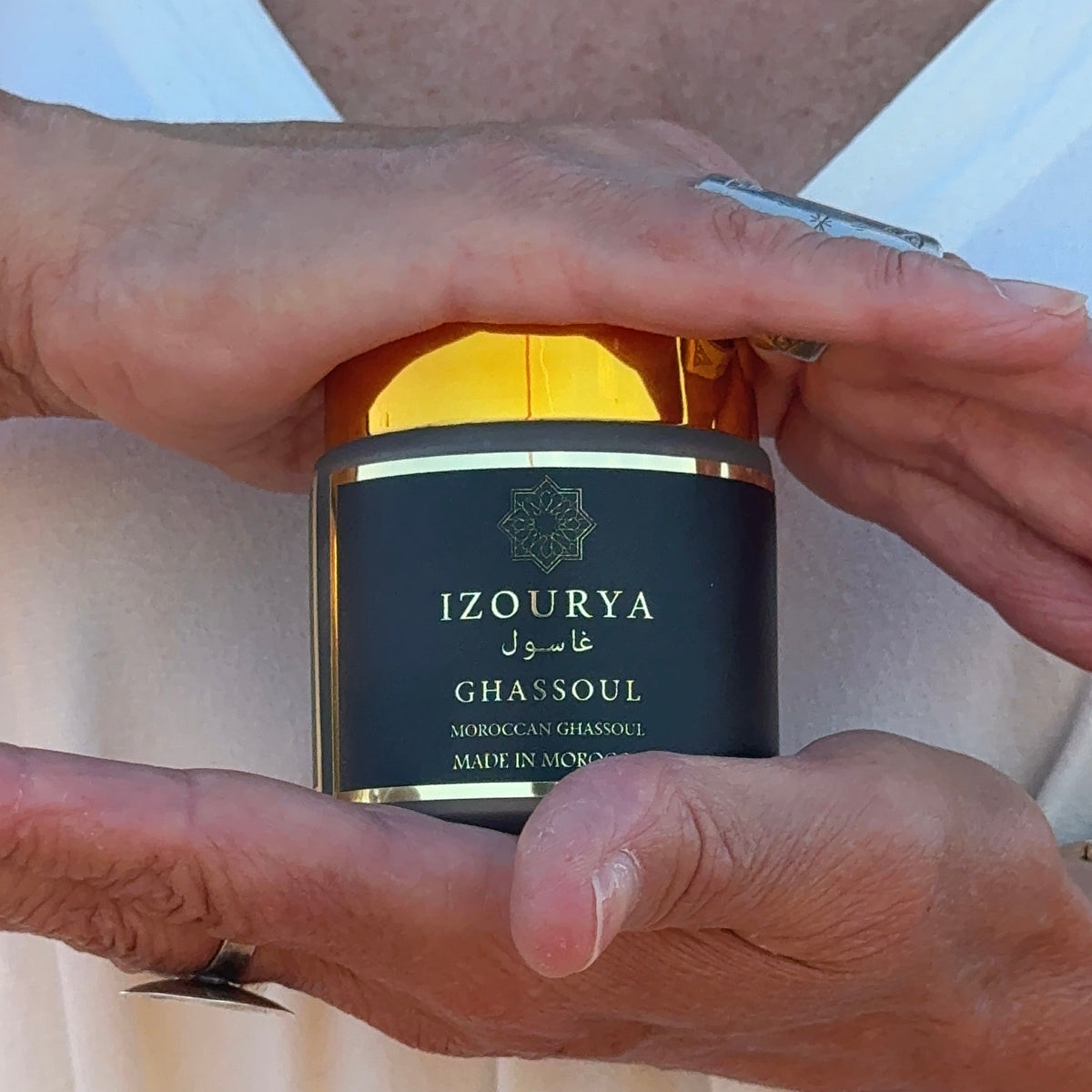 A close-up of a person holding a jar of IZOURYA Ghassoul face clay mask. The jar has a black label with gold lettering and a shiny gold lid, held in hands wearing silver rings against a cream-colored dress.