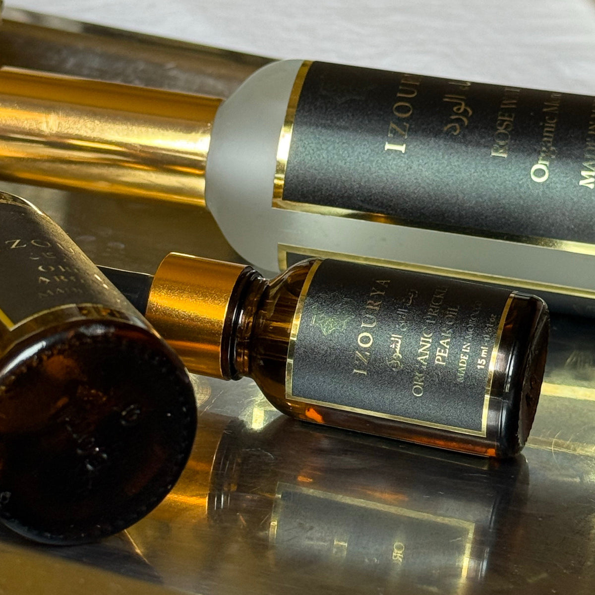Three cosmetic bottles from the brand izourya, including an amber glass body wash bottle, a frosted bottle, and an amber spray bottle, arranged on a gold tray with a white fabric background.