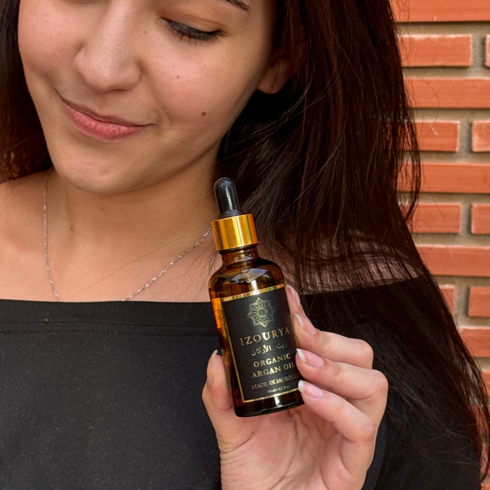 A woman holding a small amber glass dropper bottle labeled 'izourya Organic argan oil' against a brick wall background.