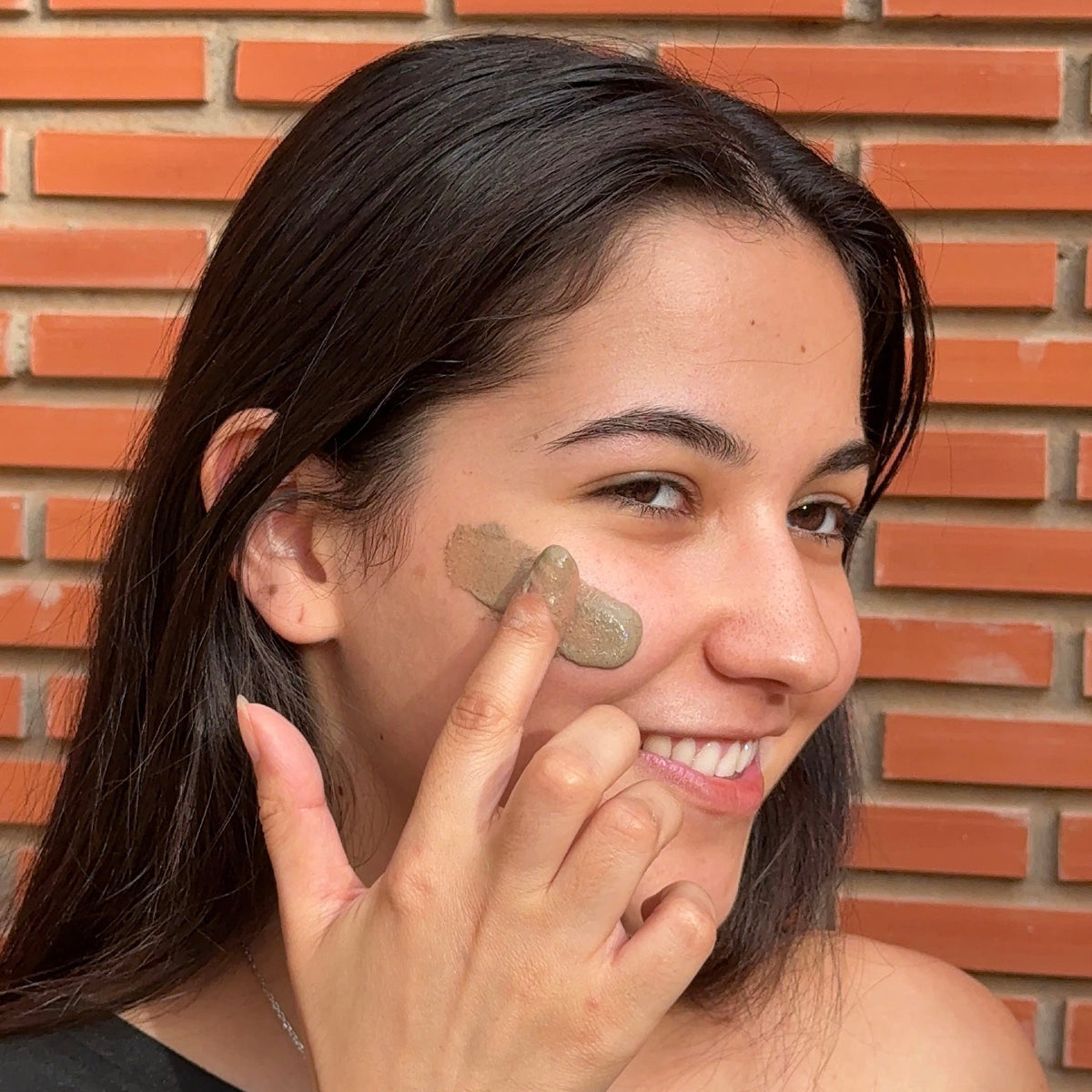 A woman with dark hair applies a beige makeup product, likely a face powder or bronzer, to her cheek in front of a red brick wall.