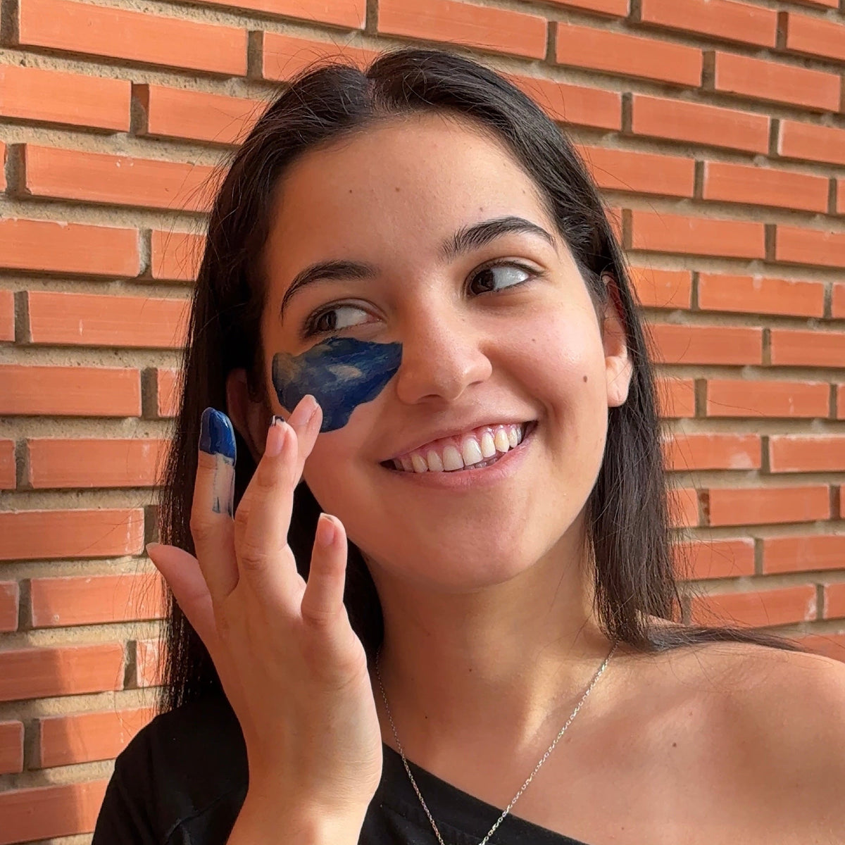 A woman with dark hair smiles while applying a blue gel or cream to her cheek, holding a small blue container. She is wearing a black off-the-shoulder top and standing against a red-orange brick wall.