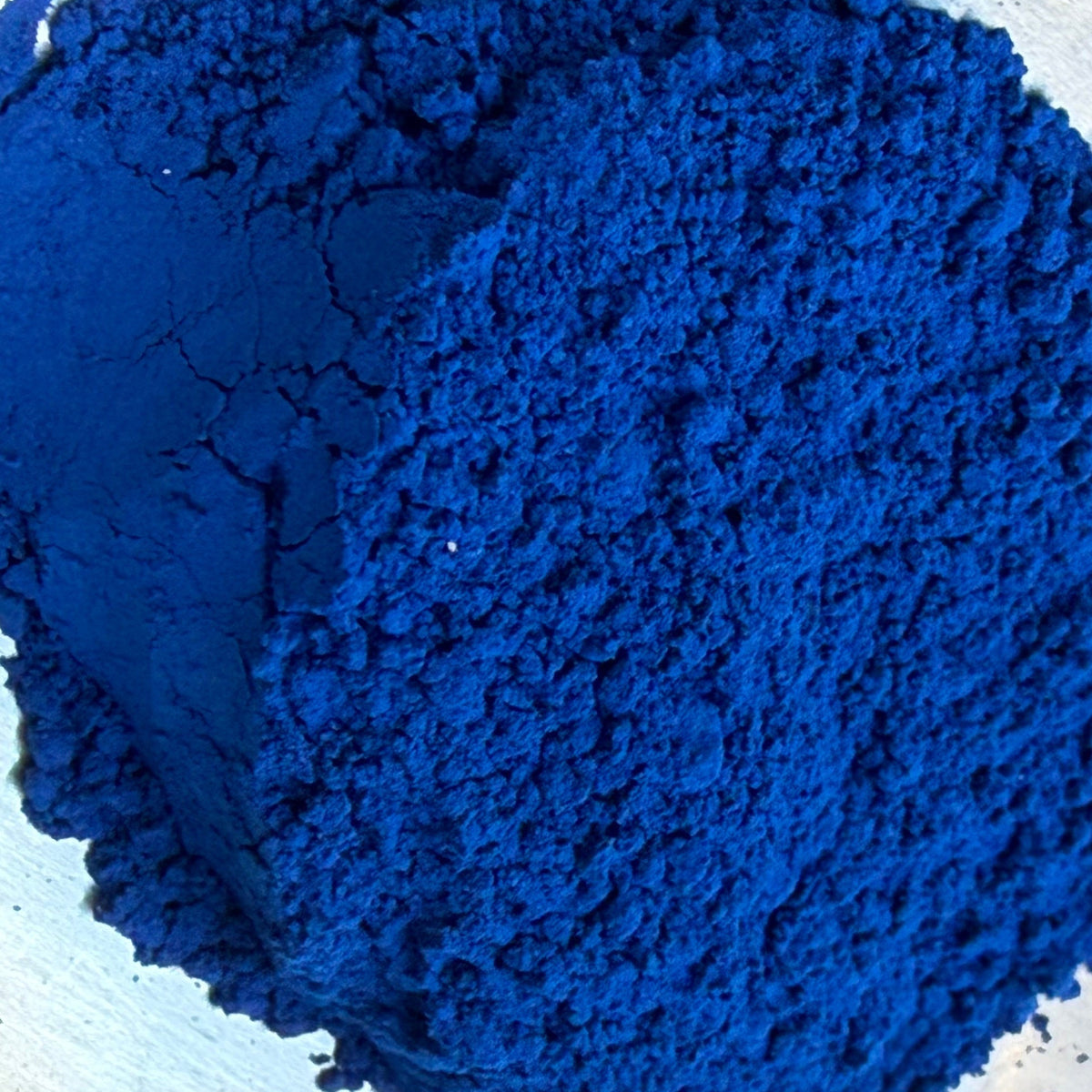 A close-up of bright cobalt blue powder piled in a silver metal spoon against a dark textured background.