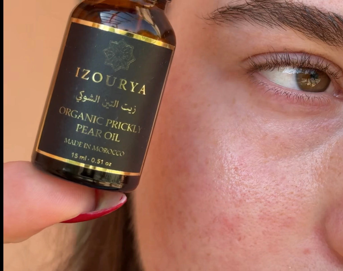 A close-up of a person's face , holding a small amber bottle of IZOURYA Organic Prickly Pear Oil next to their cheek.