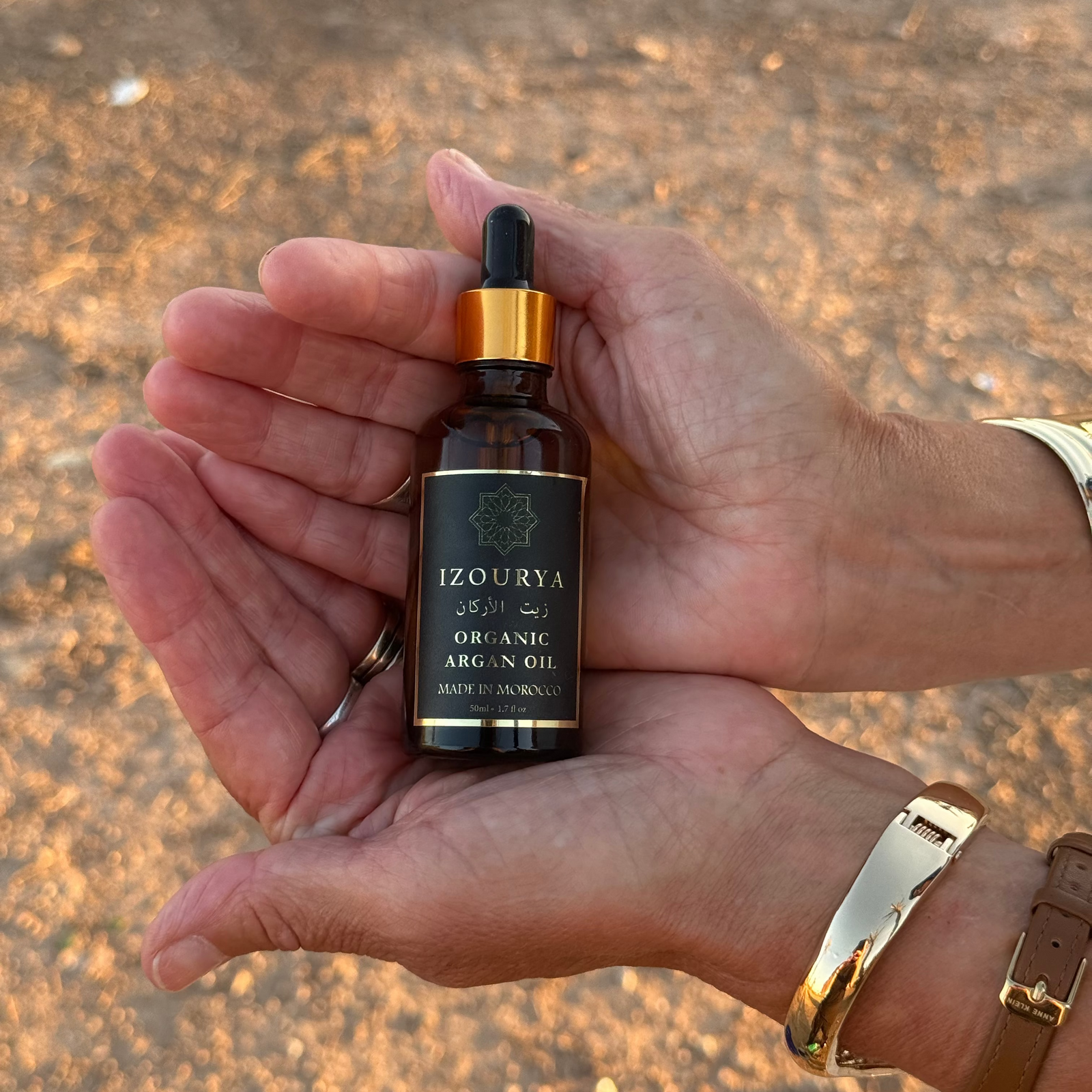 A hand holding a small amber dropper bottle of IZOURYA Organic Argan Oil against a blurred sandy outdoor background.