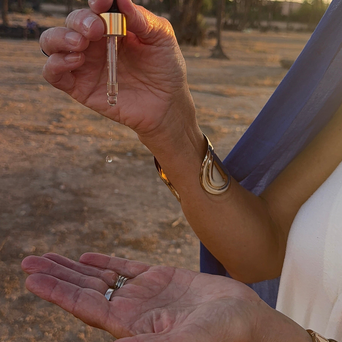 A close-up of a person outdoors at sunset holding a small glass dropper bottle with a gold cap, with a clear liquid droplet falling. The person's hands are visible, along with a white top, gold bracelets, and a blue scarf, with palm trees and dry ground in the background.