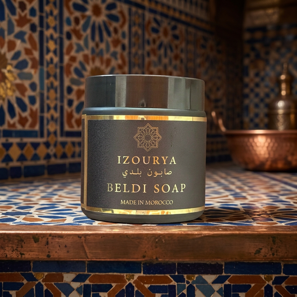 A dark gray jar of IZOURYA BELDI SOAP with gold lettering, placed on a wooden surface against a background of blue and gold Moroccan patterned tiles.