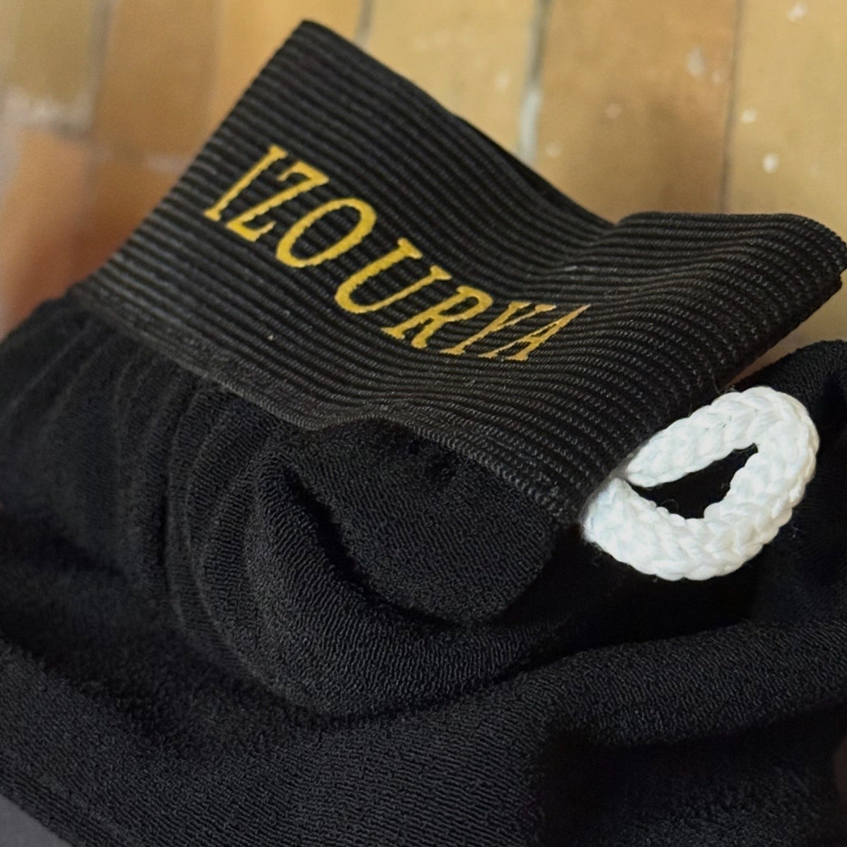 A black ribbed knit accessory with gold 'IZOURYA' lettering on the cuff, folded on a dark surface against a tan tiled background.